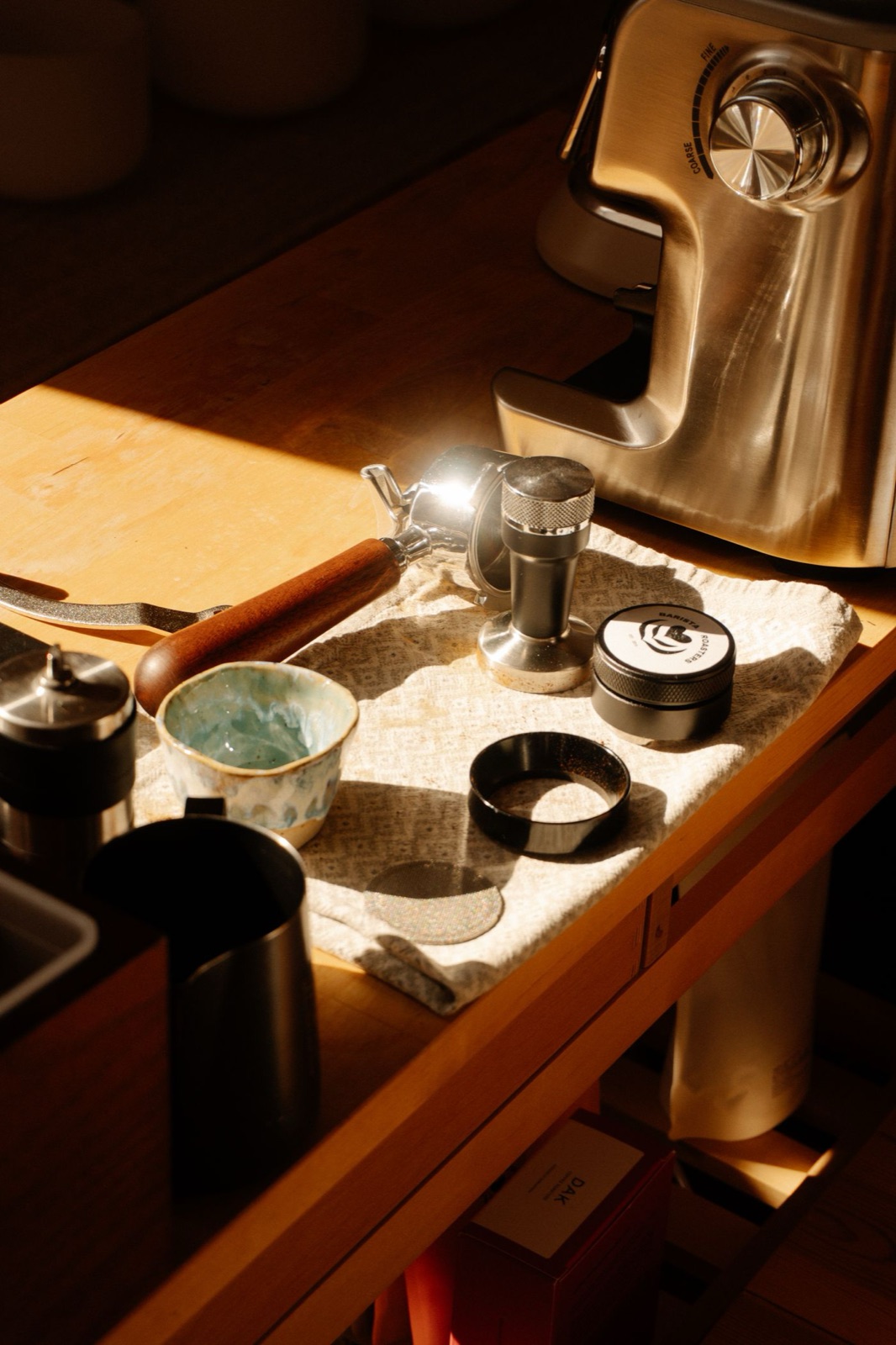 Bancada do barista com porta-filtro, tamper, moinho e jarra sob luz de janela